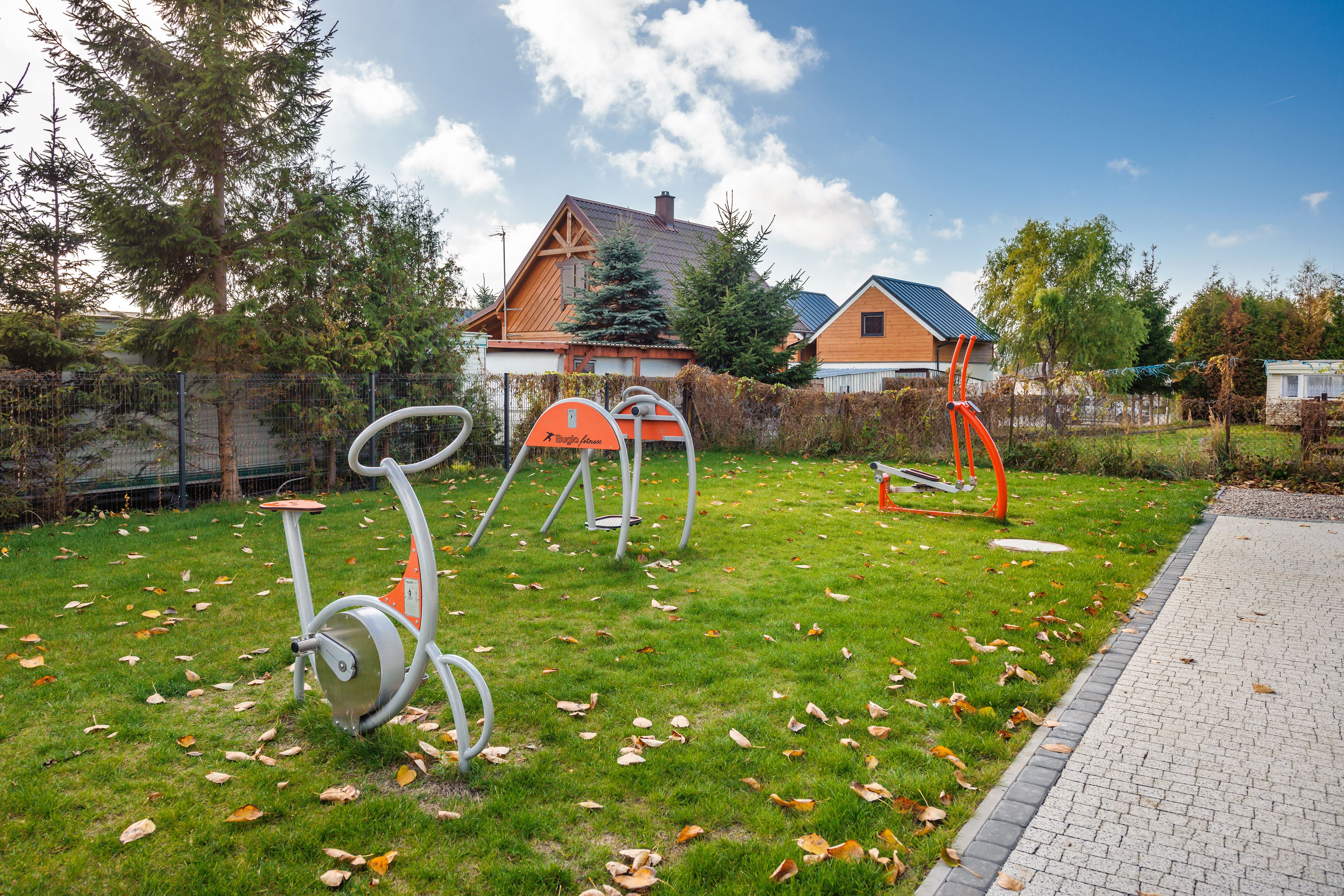 An outdoor exercise area with stationary bikes and a slide, surrounded by a grassy field and a brick pathway.