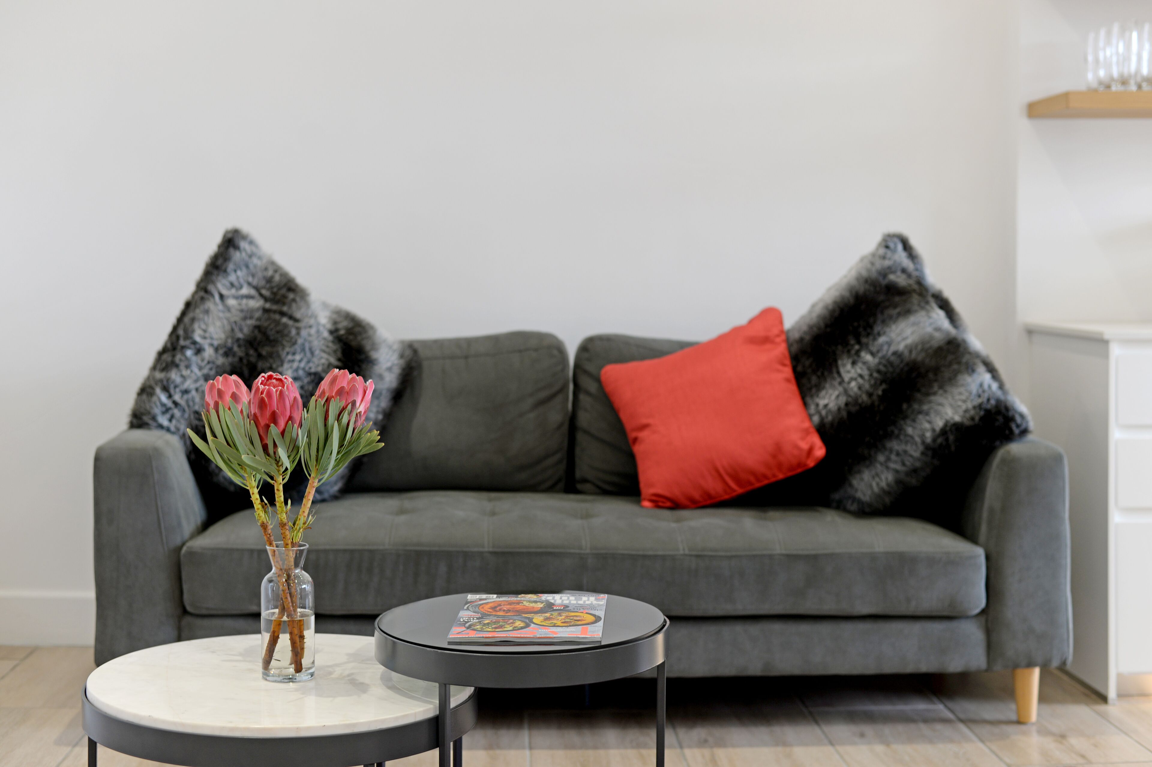 A modern living room with a grey sofa, two black and white pillows, a red pillow, a vase with pink flowers, and a magazine on a small round table.