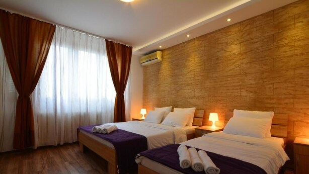 Habitación de hotel con dos camas, suelo de madera, pared con textura de piedra y una ventana con cortinas marrones.