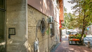 Fachada de edificio con unidades de aire acondicionado, una reja metálica y una acera bordeada de árboles.