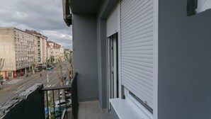 Een balkon met uitzicht op een stadstraat, een gebouw met een raam met witte luiken, en een heldere lucht.