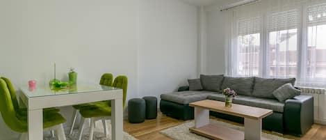 A modern living room with a glass-top dining table, green chairs, a grey sofa, and a white coffee table.
