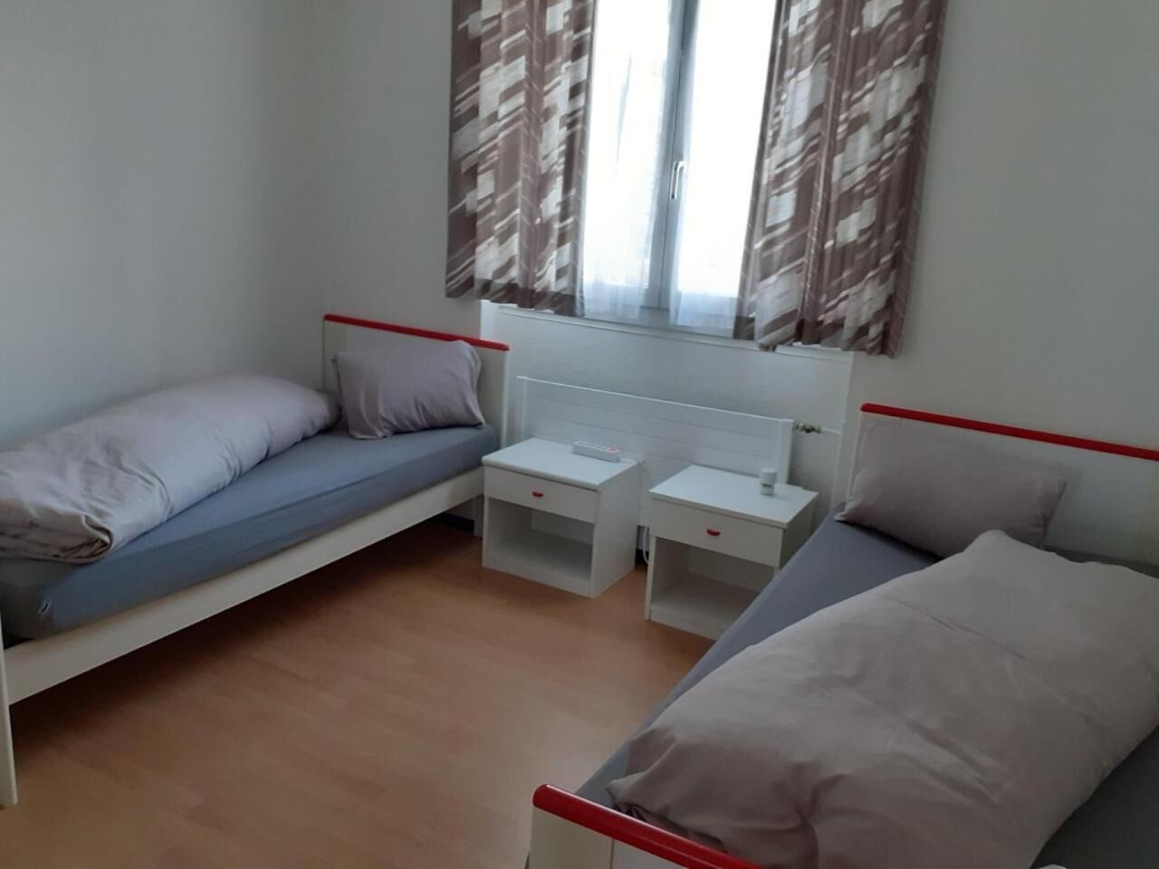 Une petite chambre propre, avec deux lits simples, une petite table et une fenêtre avec des rideaux.