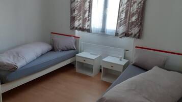 Une petite chambre propre, avec deux lits simples, une petite table et une fenêtre avec des rideaux.