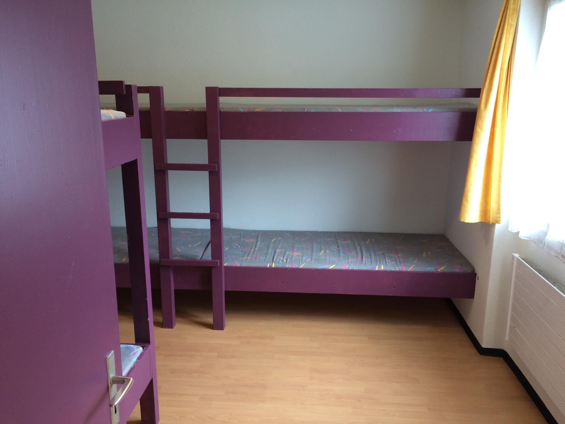 A bunk bed room with a ladder, wooden flooring, and a window with curtains.