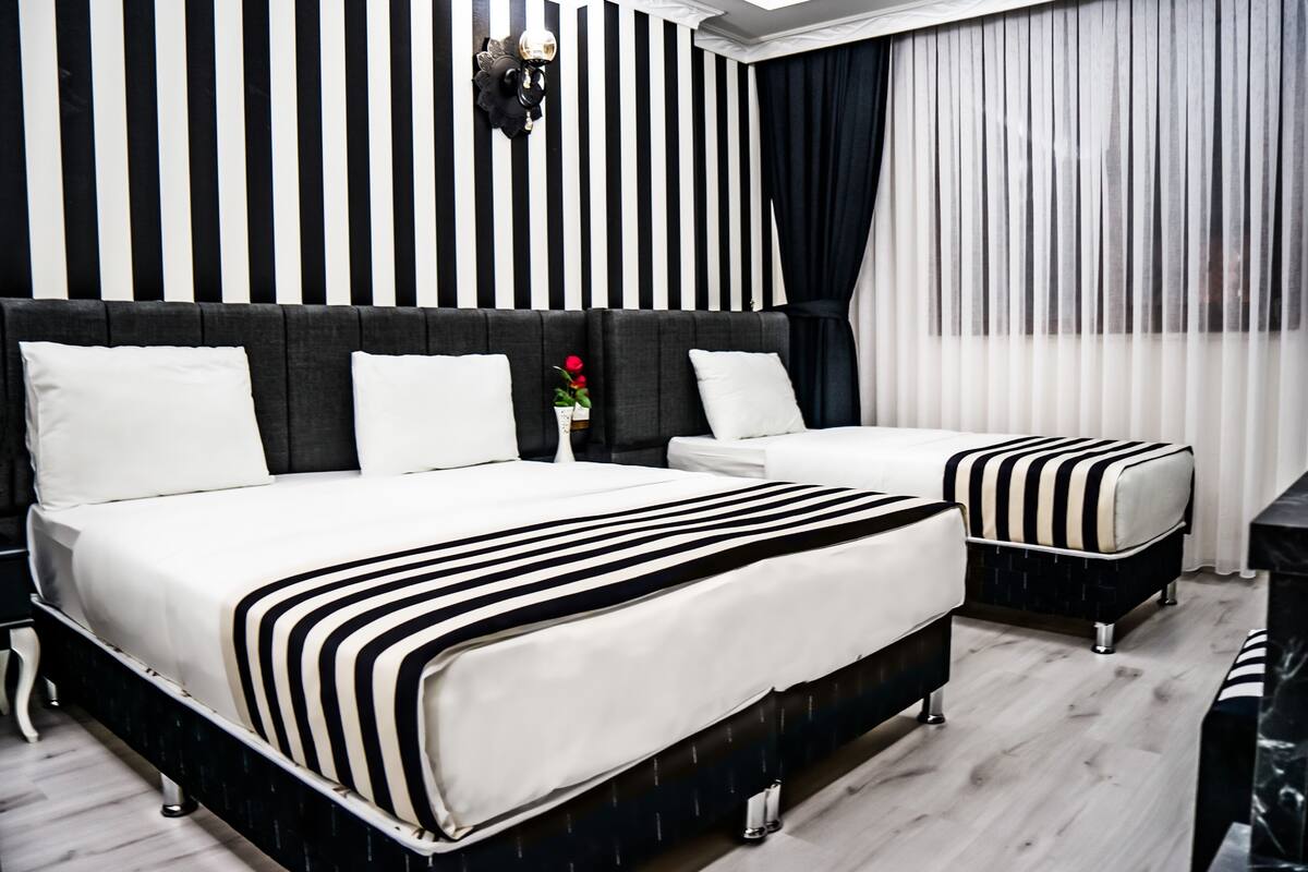 Une chambre d’hôtel avec un grand lit, un lit plus petit, une table de chevet et un pouf rayé noir et blanc.
