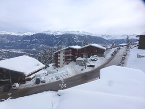 A snow-covered mountain town with a road lined with parked cars and buildings, including a multi-story hotel with red and white exterior. - Appartement Emeraude 615 (Ayent)