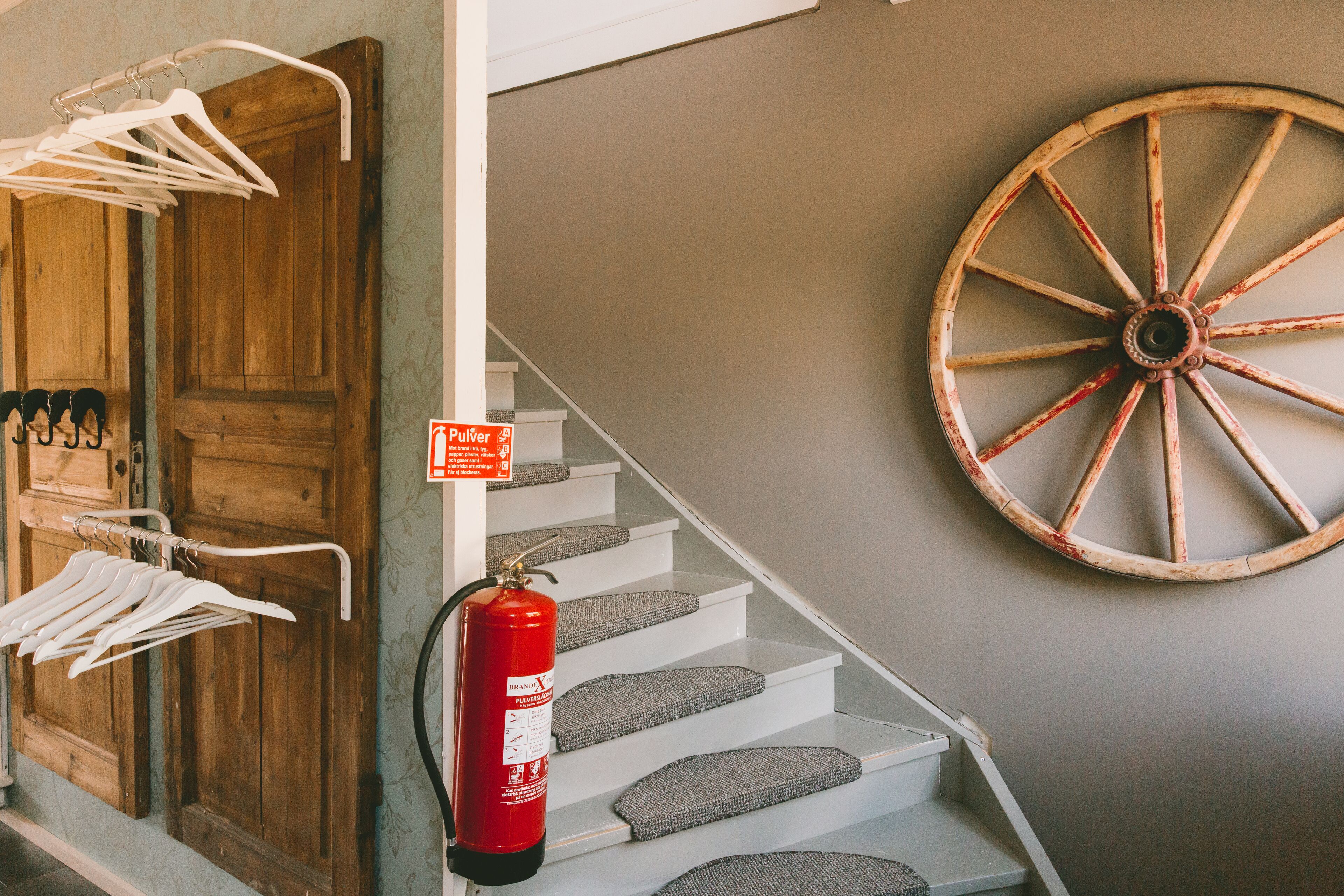 Un escalier avec des marches recouvertes de moquette grise, un extincteur fixé au mur et une porte en bois avec un porte-manteau.