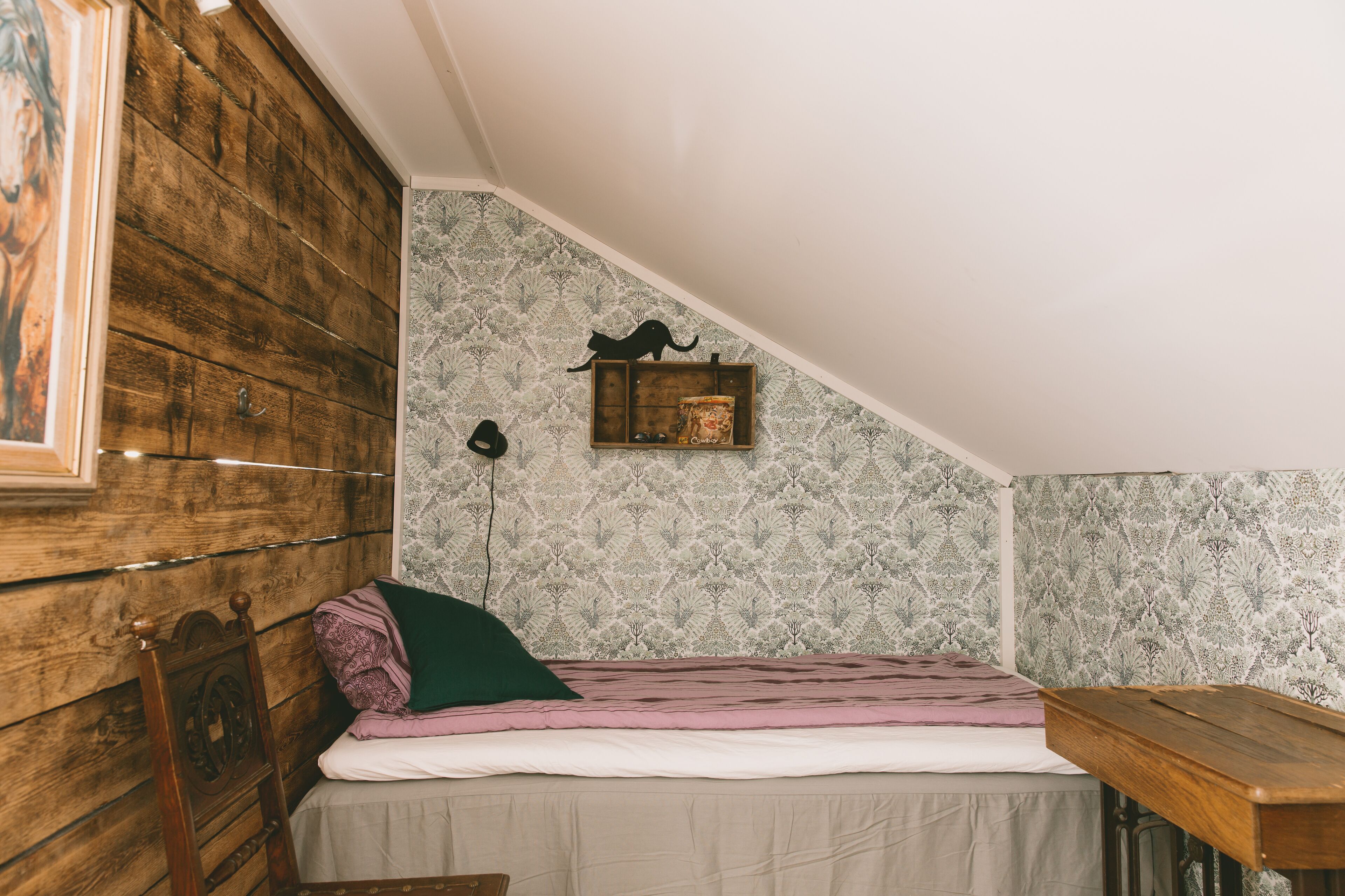 Ein kleiner, gemütlicher Raum mit schrägem Dach, einem Holzkopfende, einem Einzelbett mit rosa-weißer Streifenoptik, einer kleinen Ablage mit einem dekorativen Gegenstand und strukturierten Tapeten.