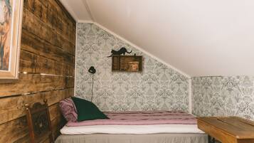 Ein kleiner, gemütlicher Raum mit schrägem Dach, einem Holzkopfende, einem Einzelbett mit rosa-weißer Streifenoptik, einer kleinen Ablage mit einem dekorativen Gegenstand und strukturierten Tapeten.