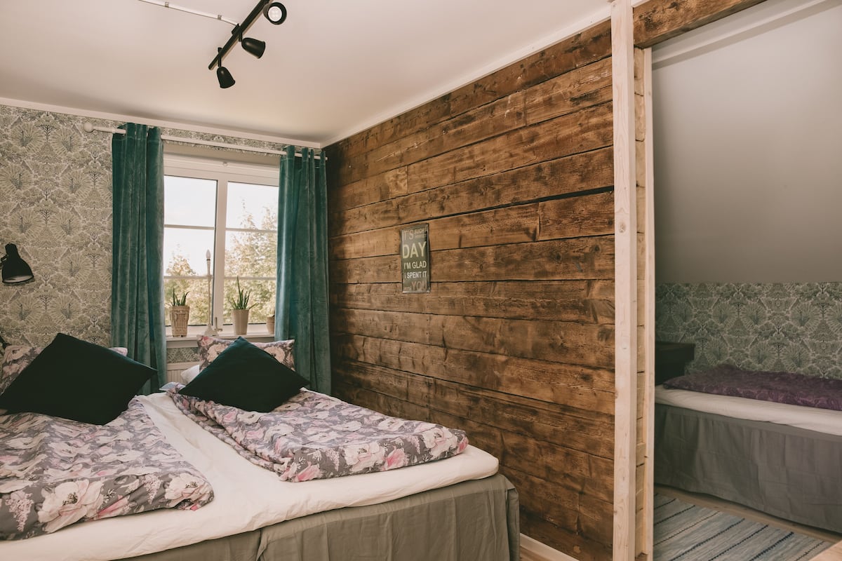 A bedroom with a wooden wall, a window with curtains, a bed with floral bedding, and a bedside table.
