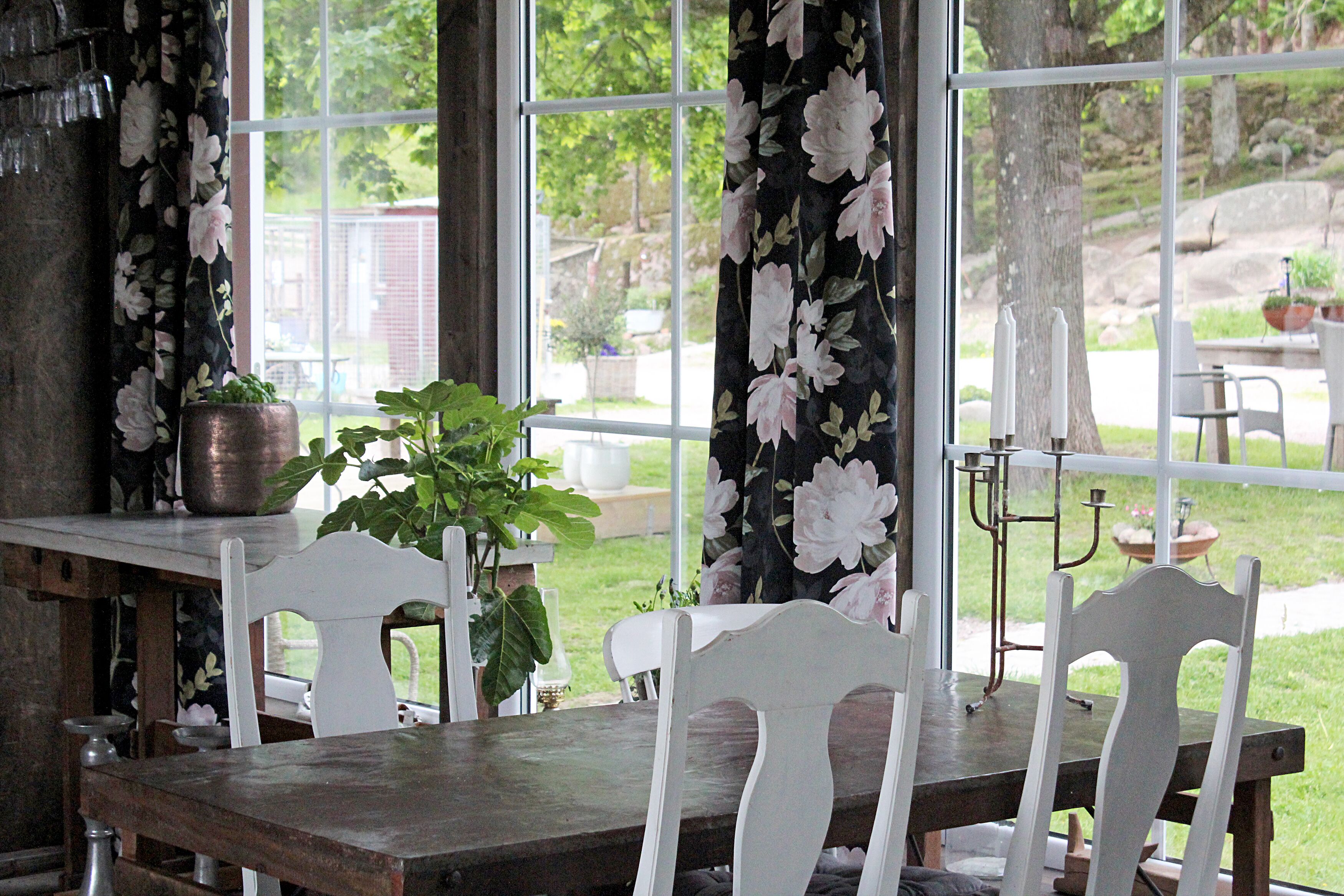En matplats med ett träbord och vita stolar, stora fönster med blommiga gardiner och utsikt över en trädgård.