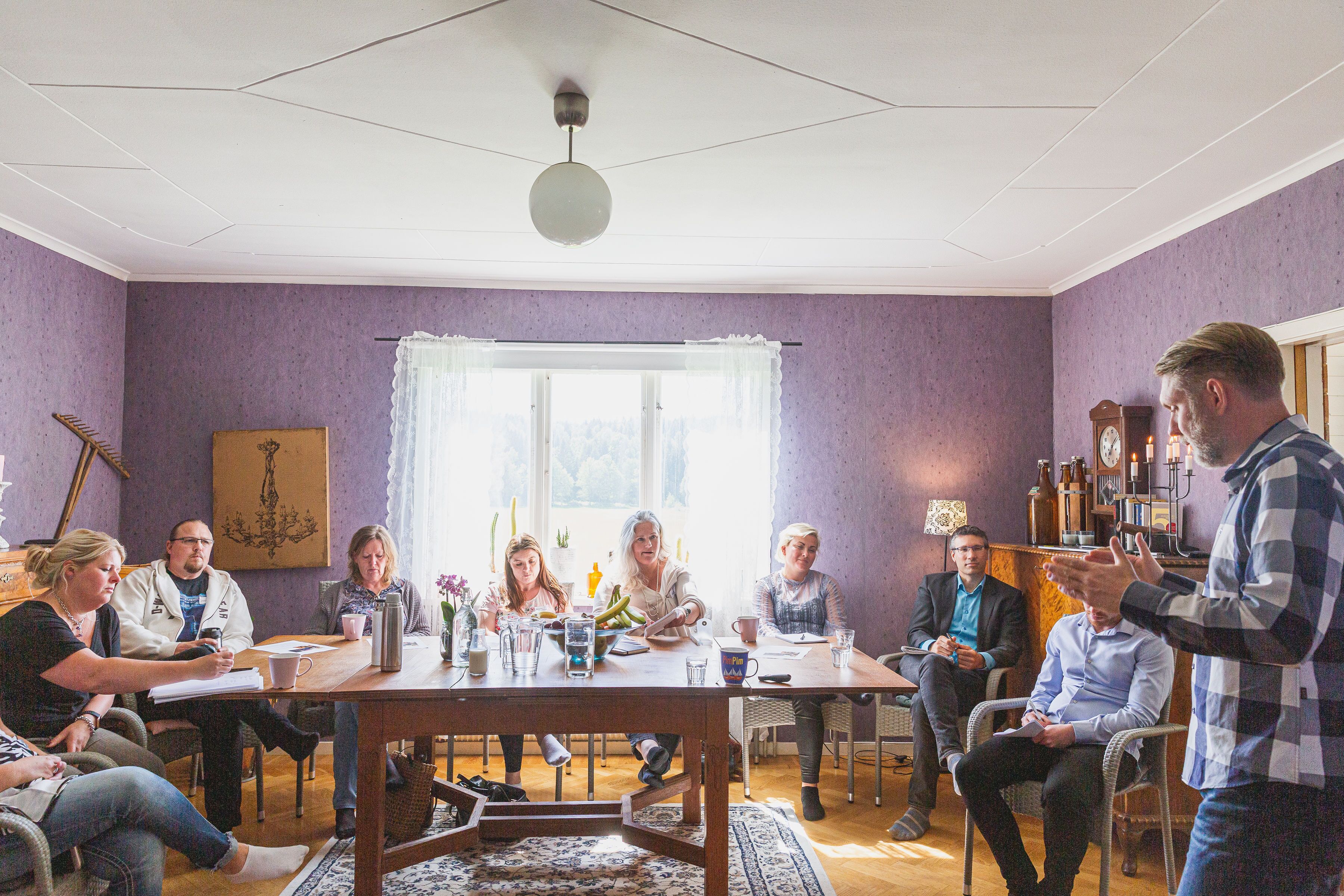 Un grupo de personas sentadas alrededor de una mesa de madera en una habitación con paredes moradas, un candelabro y un ventanal con cortinas.