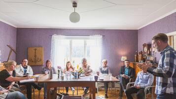 Un grupo de personas sentadas alrededor de una mesa de madera en una habitación con paredes moradas, un candelabro y un ventanal con cortinas.