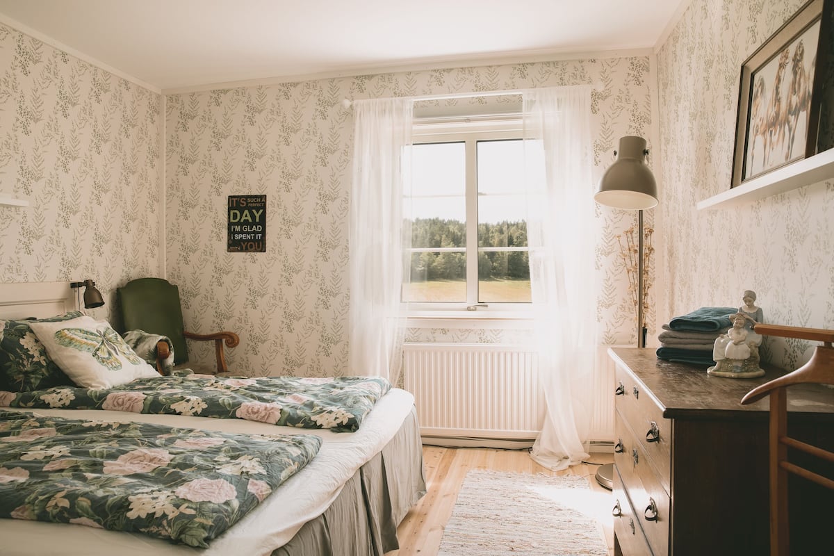 A bedroom with floral wallpaper, a bed with floral bedding, a radiator, a dresser with a lamp, and a window with a view of trees.