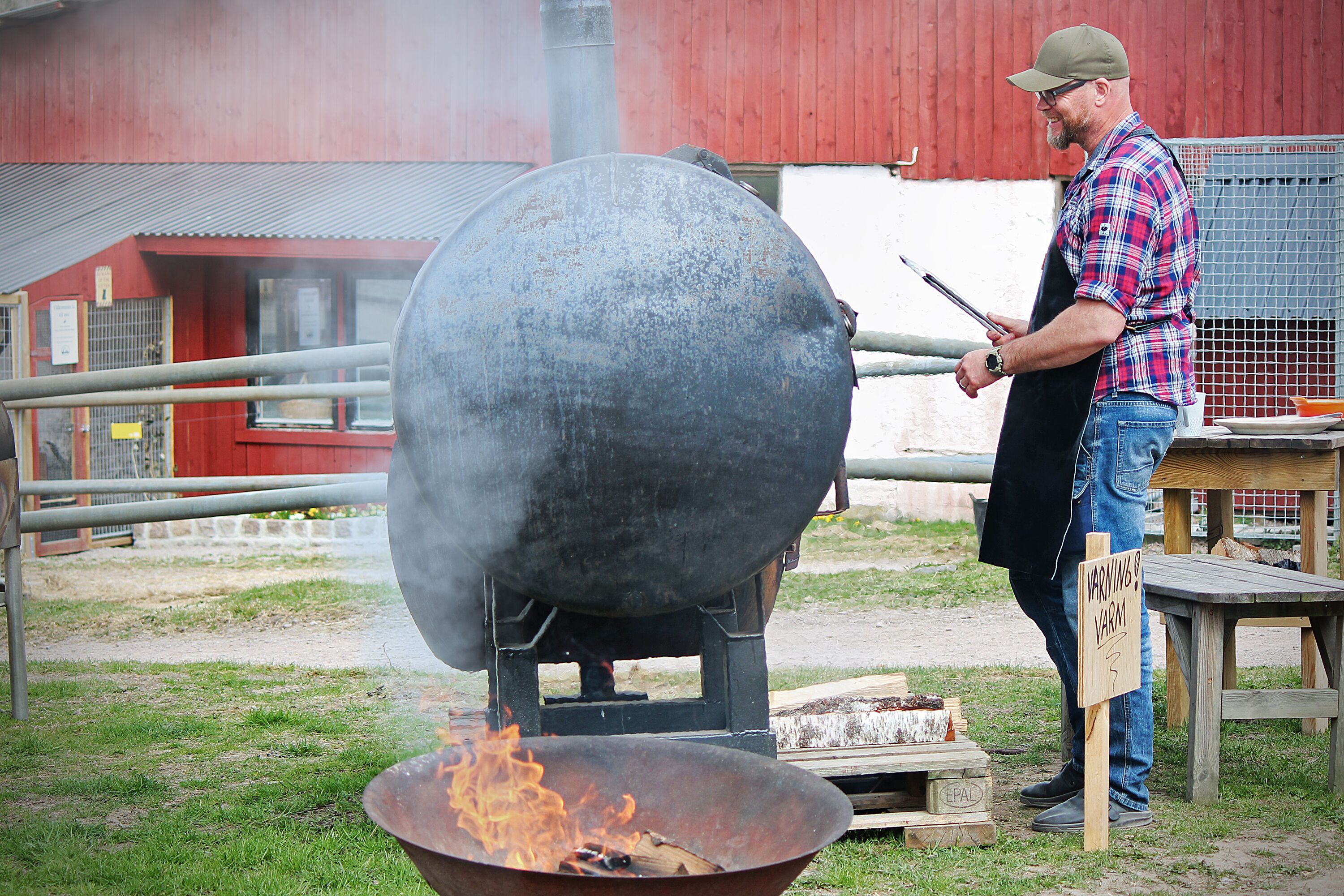 A man grilling outdoors with a large smoker and a warning sign nearby.