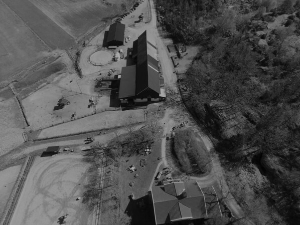 An aerial view of a rural property with a large barn, several smaller buildings, and a fenced area with a circular track. - Farmlodge (Brastad)