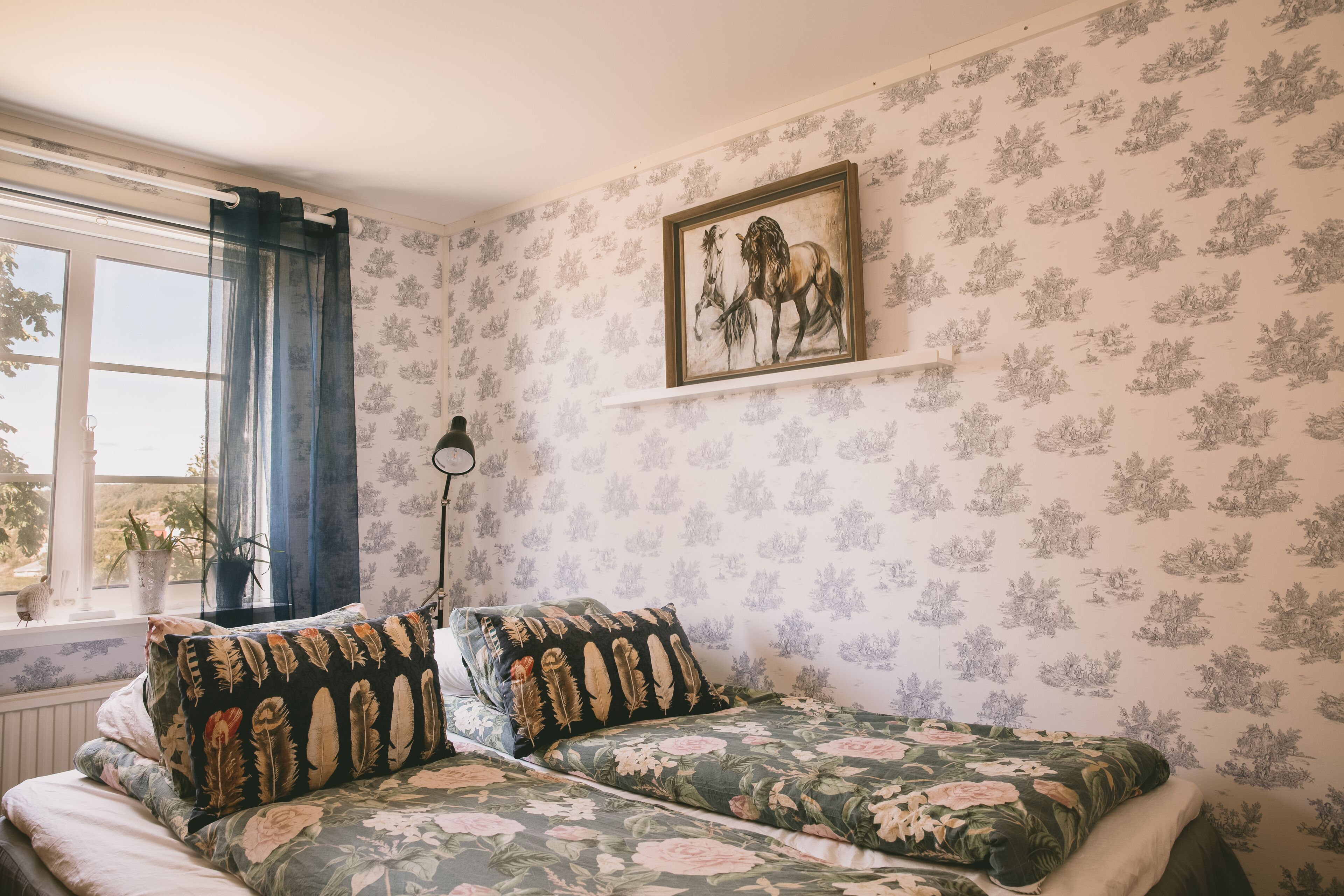 Una habitación con papel tapiz floral, una cama con ropa de cama estampada con flores, una ventana con cortinas y un cuadro en la pared.