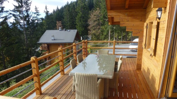 A wooden deck with a glass-top table and wicker chairs, overlooking a forested area. - Chalet Clématites (Ayent)