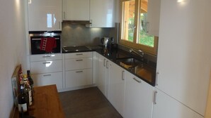 A modern kitchen with white cabinets, a black countertop, a built-in oven, and a window with a view of greenery. - Chalet Clématites (Ayent)