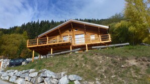 A wooden cabin with a balcony, situated on a hillside with a rocky foreground and forested background. - Chalet Clématites (Ayent)