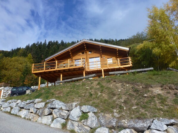 A wooden cabin with a balcony, situated on a hillside with a rocky foreground and forested background.