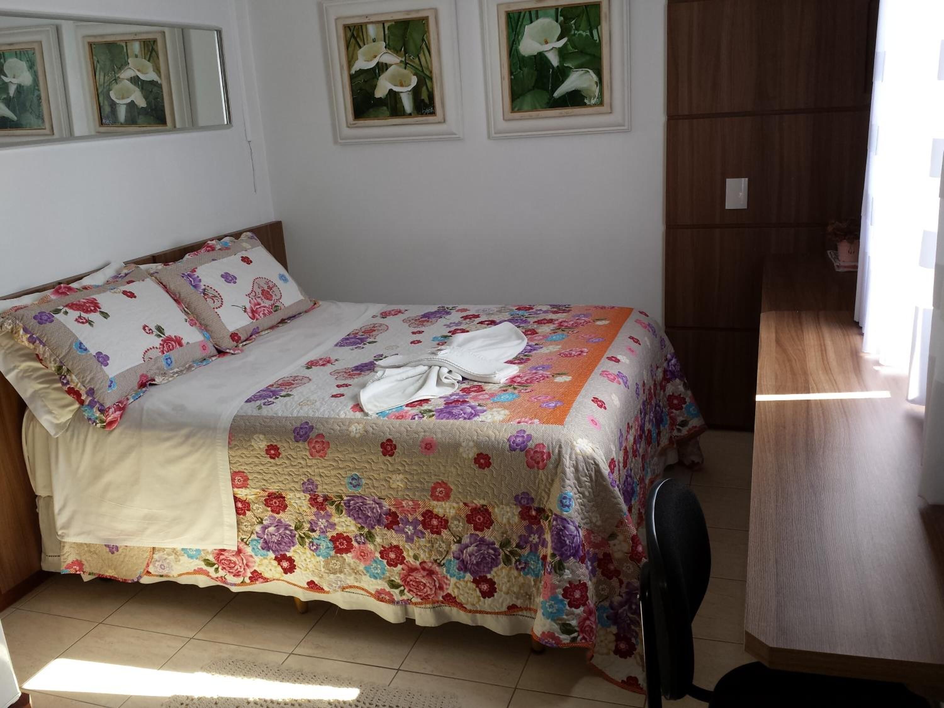 Uma cama bem arrumada com roupa de cama floral, uma mesinha de cabeceira e uma cadeira.