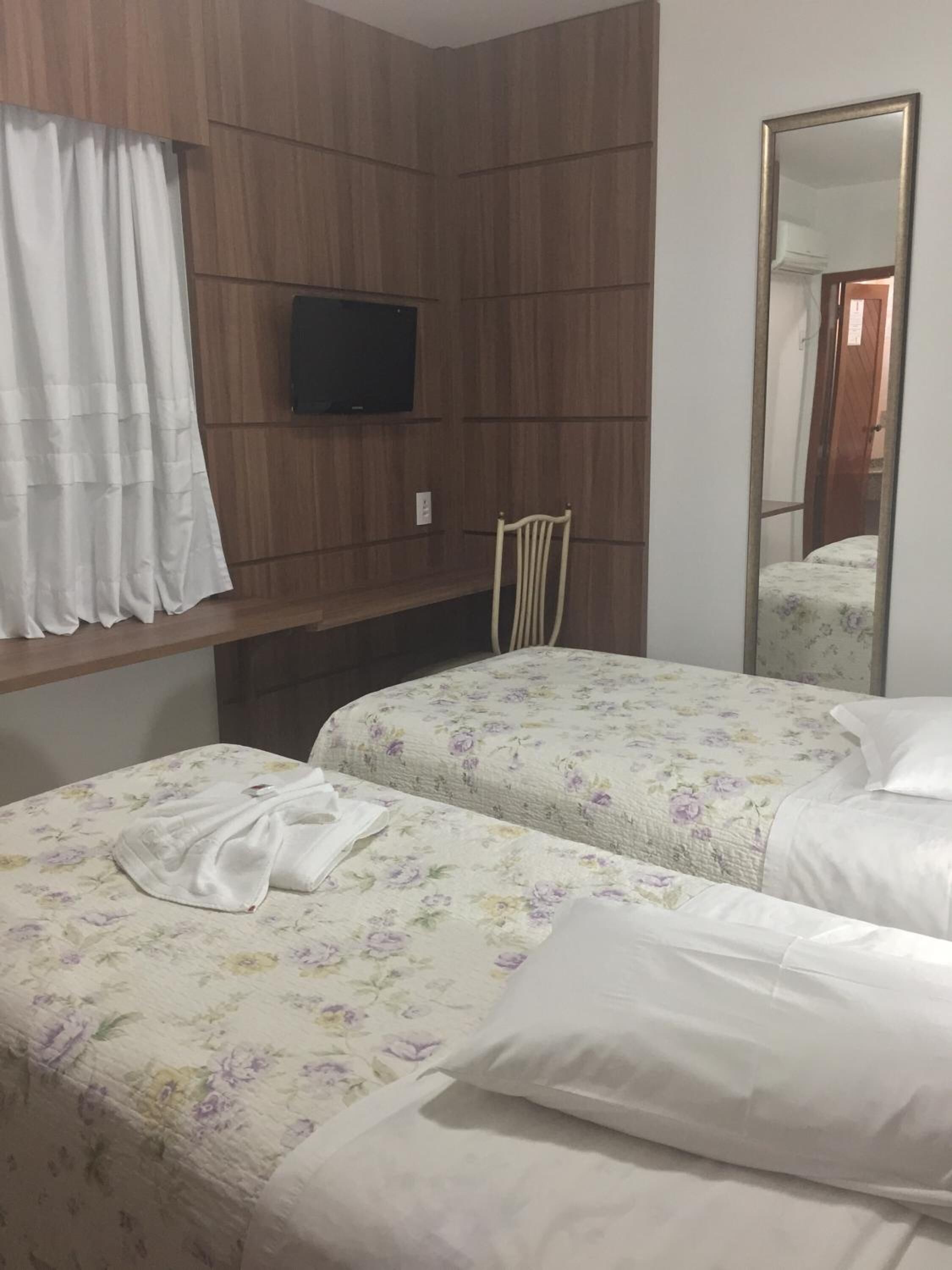 Ein Hotelzimmer mit zwei Betten, einem an der Wand befestigten Fernseher, einer Holzpaneel-Wandgestaltung, einem Spiegel und einem fenster mit Vorhängen.