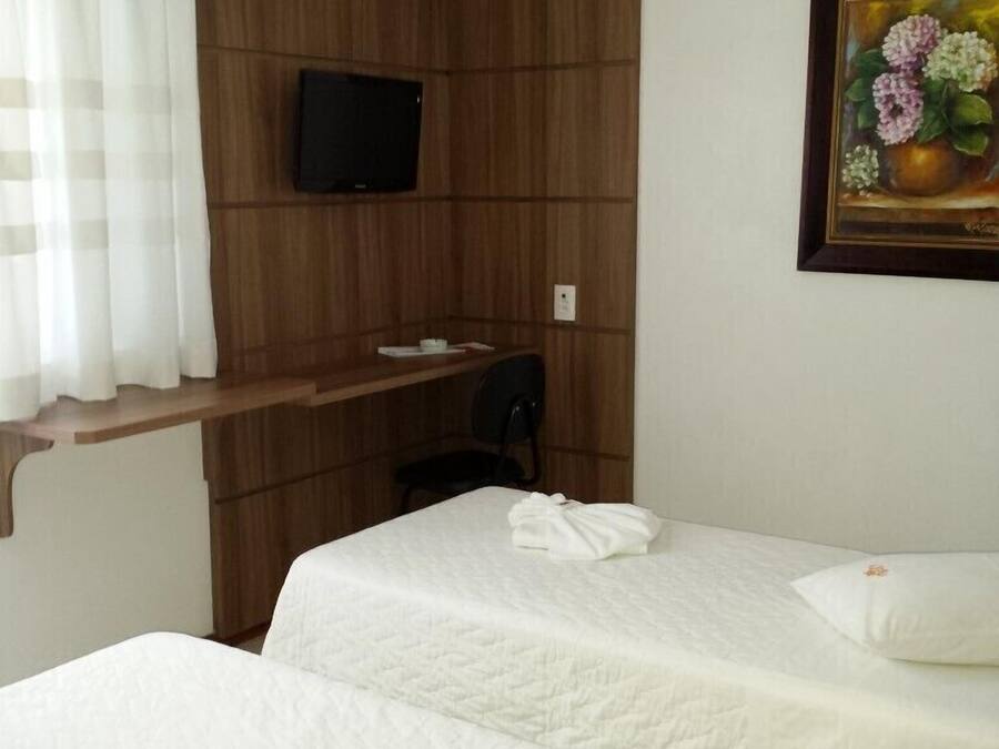 A hotel room with two beds, a TV mounted on the wall, a painting of flowers, and a window with curtains.