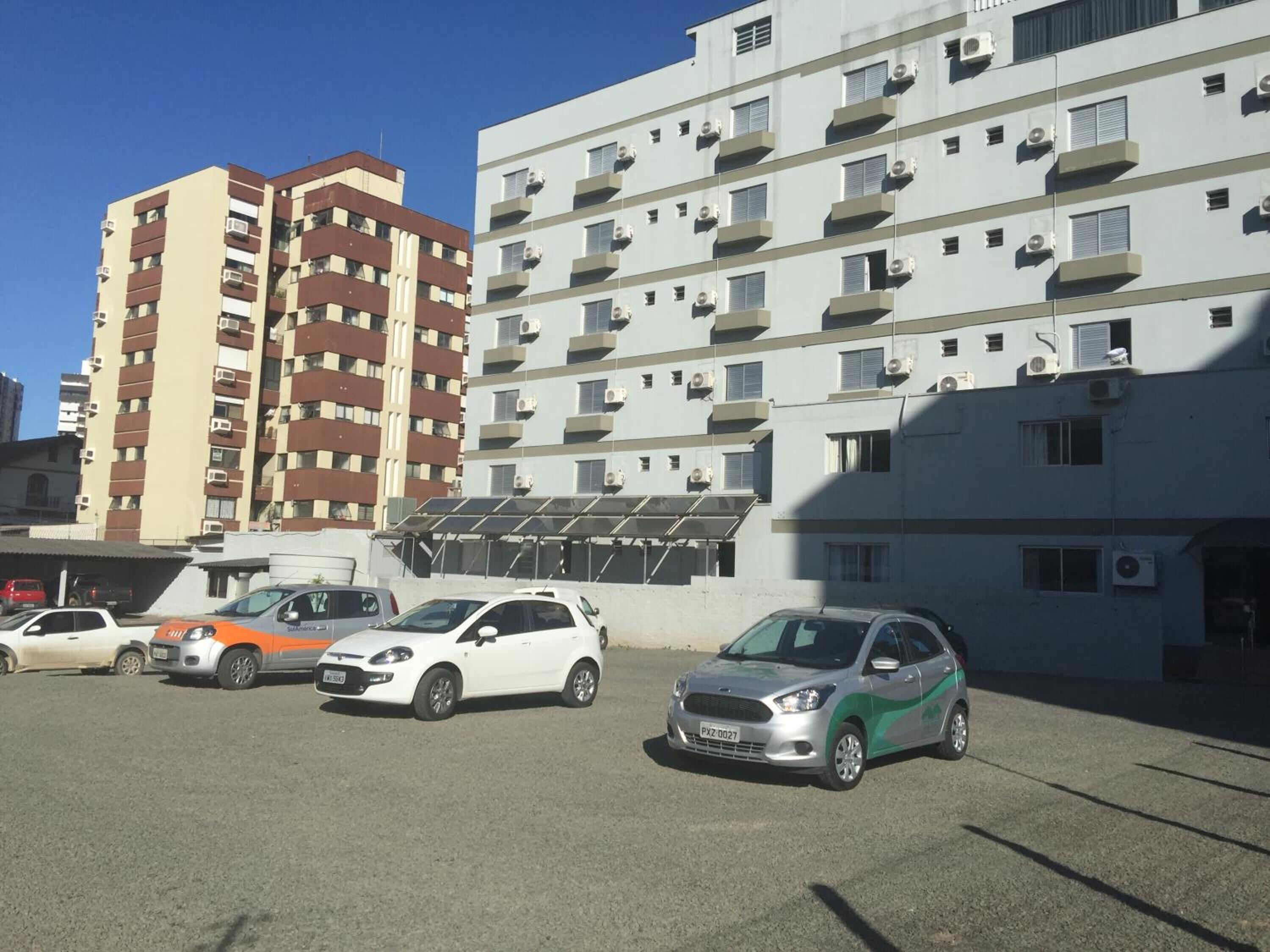 A parking lot with several cars, a multi-story building with balconies and air conditioning units, and a clear blue sky.
