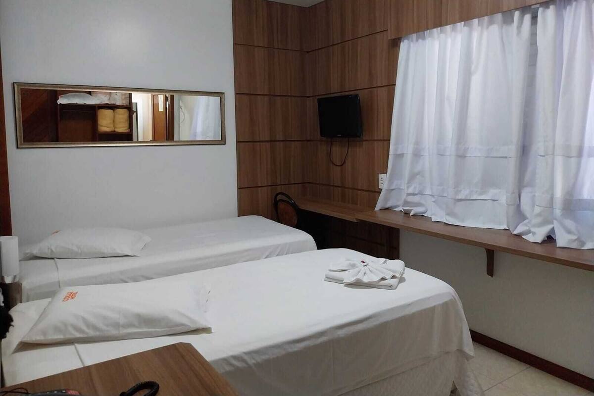 A hotel room with two single beds, a wooden panel wall, a TV mounted above a desk, and a window with white curtains.