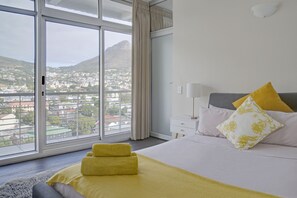 Ett hotellrum med ett stort fönster som vetter mot staden, en snyggt bäddad säng med gula handdukar och ett nattduksbord med en lampa.