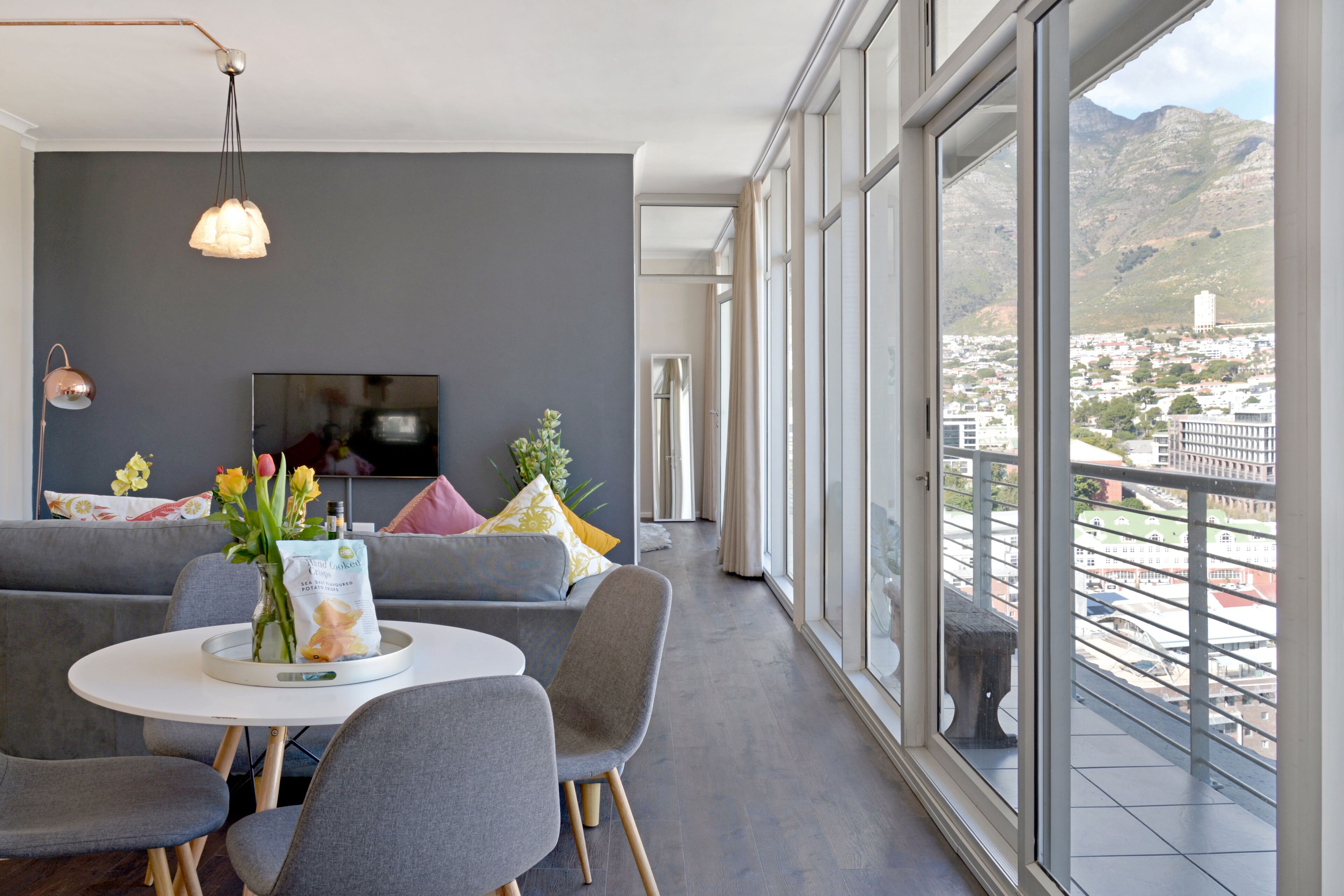 Un salon moderne avec une grande fenêtre, des canapés gris, une table ronde avec des chaises, un téléviseur et un vase rempli de fleurs.