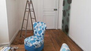 Una habitación con una silla azul con lunares, una escalera de madera, una puerta blanca y una puerta de vidrio con cortina estampada.