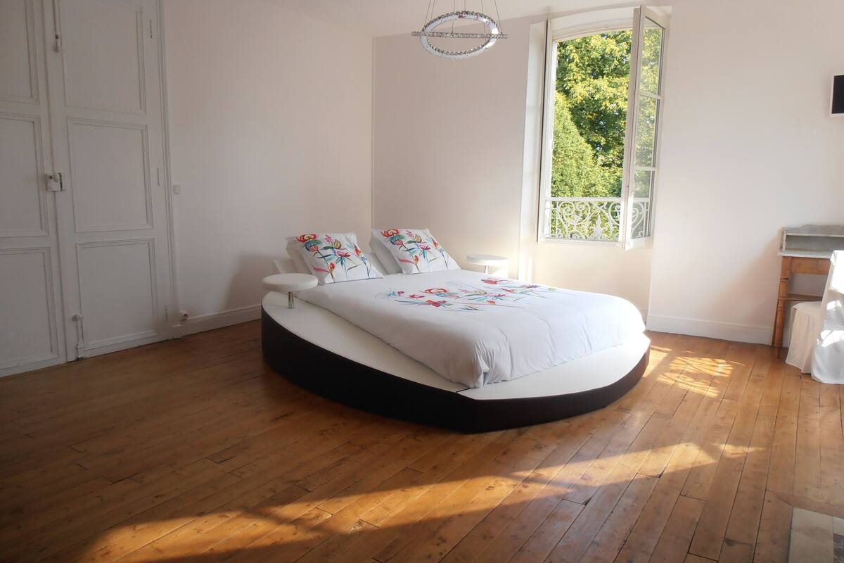 A bedroom with a curved bed, white walls, a wooden floor, a window with a view of trees, and a hanging light fixture.