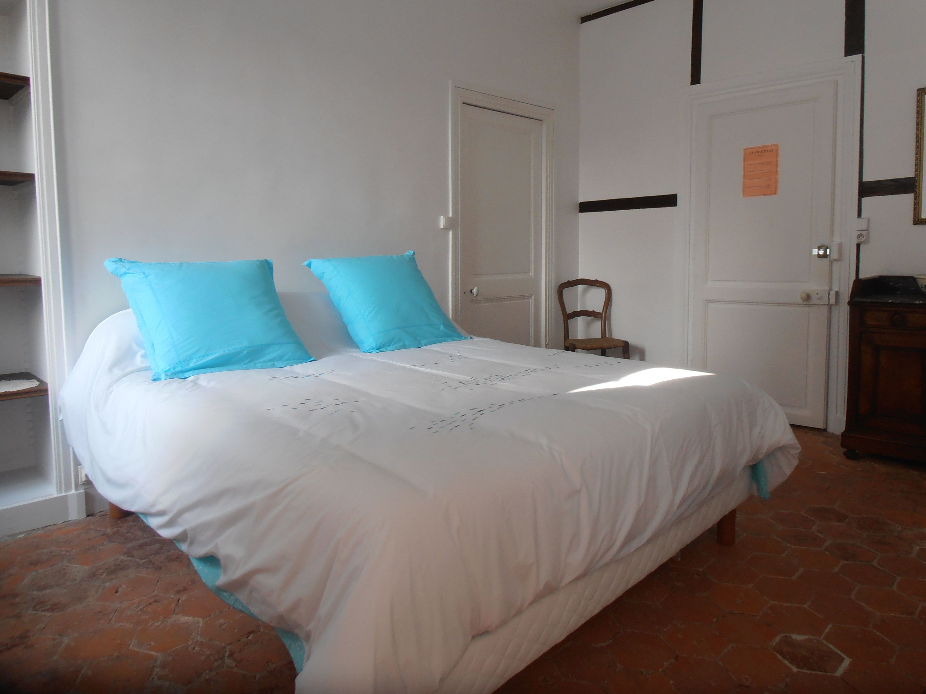 A bed with white bedding and two blue pillows, a wooden chair, and a white door with a small sign.