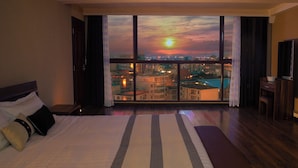 Ein Hotelzimmer mit einem großen Bett, Blick auf eine Stadtlandschaft bei Sonnenuntergang und einem Balkon mit Pool.