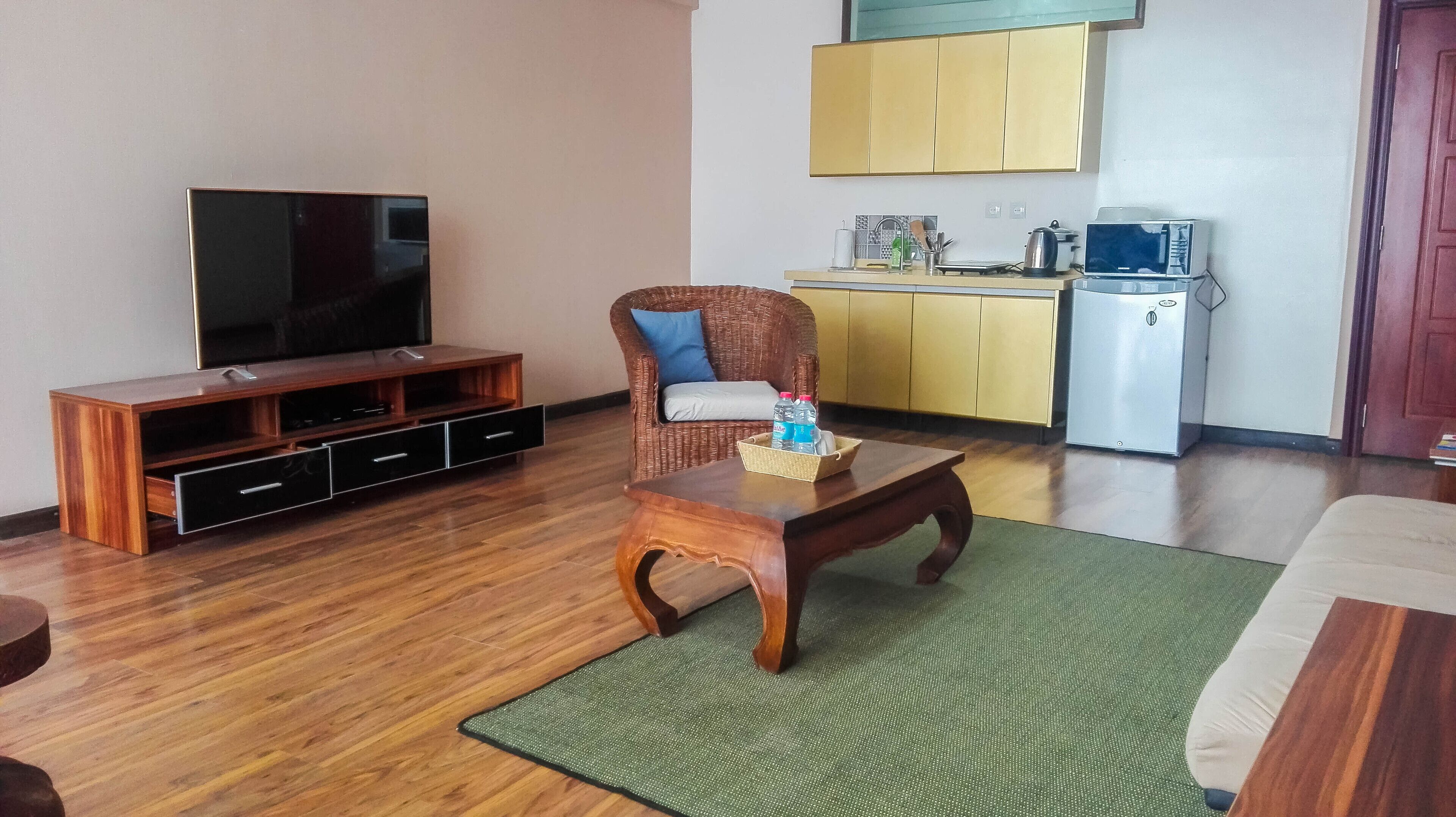 A living room with a wooden entertainment unit, a television, a wicker chair, a coffee table with water bottles, a kitchen area with yellow cabinets, and a refrigerator.