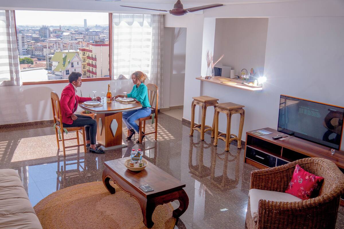 Ein Wohnzimmer mit Esstisch, zwei Stühlen, einer Couch, einem Fernseher und Blick auf die Stadt durch große Fenster.