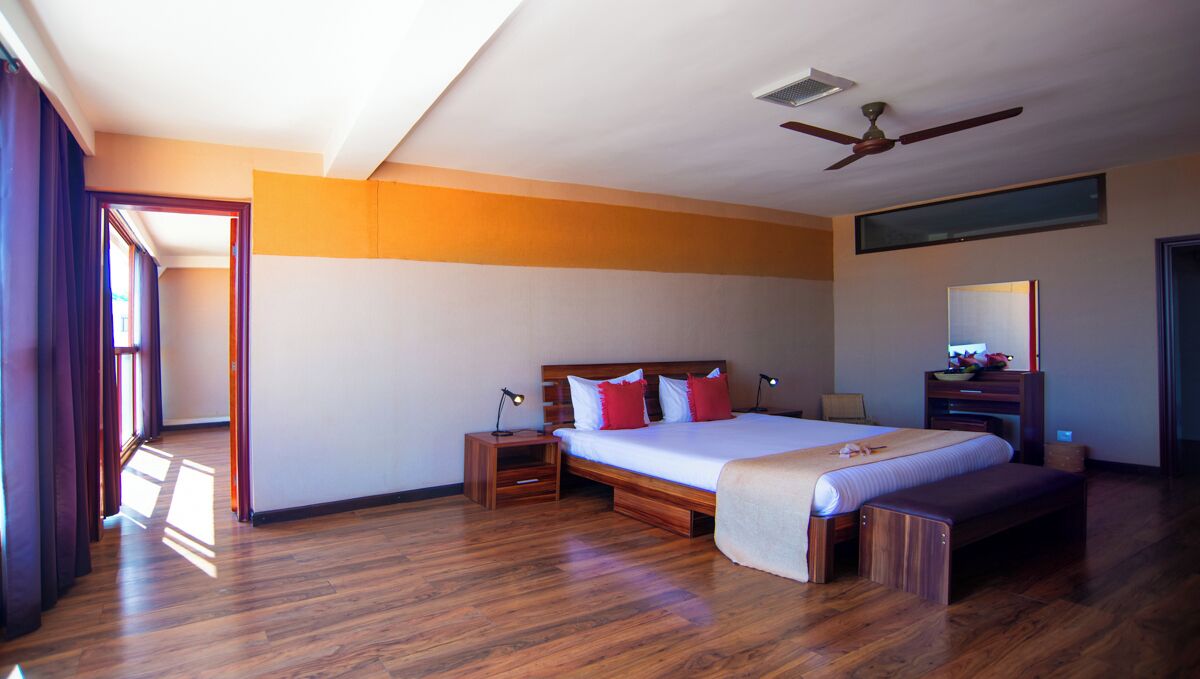 Une chambre avec un plancher en bois, un lit avec une literie rouge et blanche, une table de chevet, un banc, un ventilateur de plafond et une fenêtre.