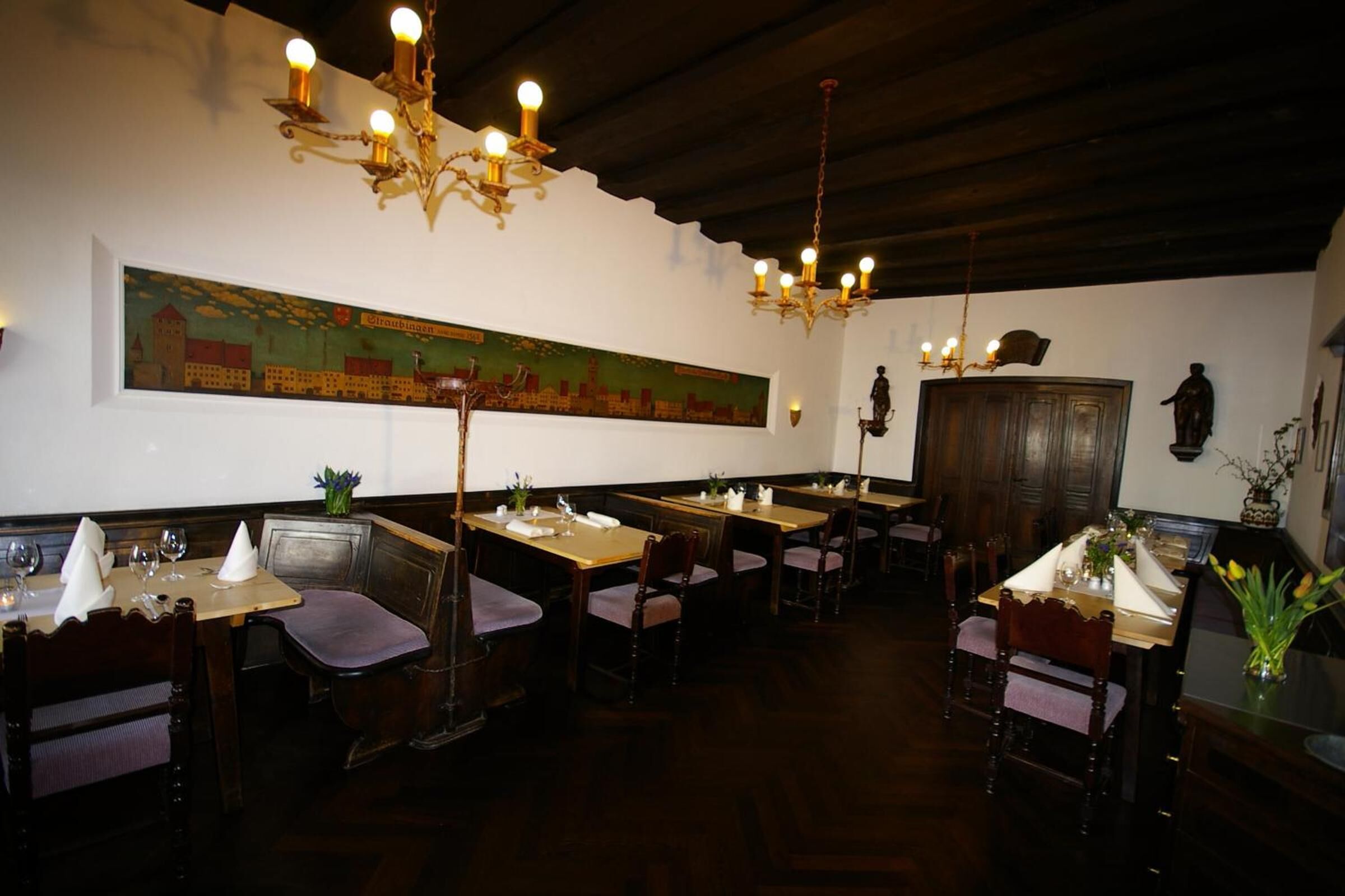 A restaurant with wooden tables and chairs, a large painting on the wall, and chandeliers.