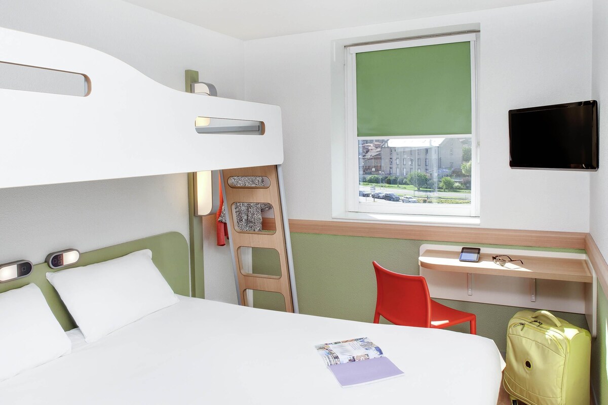 A hotel room with a bunk bed, a desk with a chair, a suitcase, and a TV on the wall.
