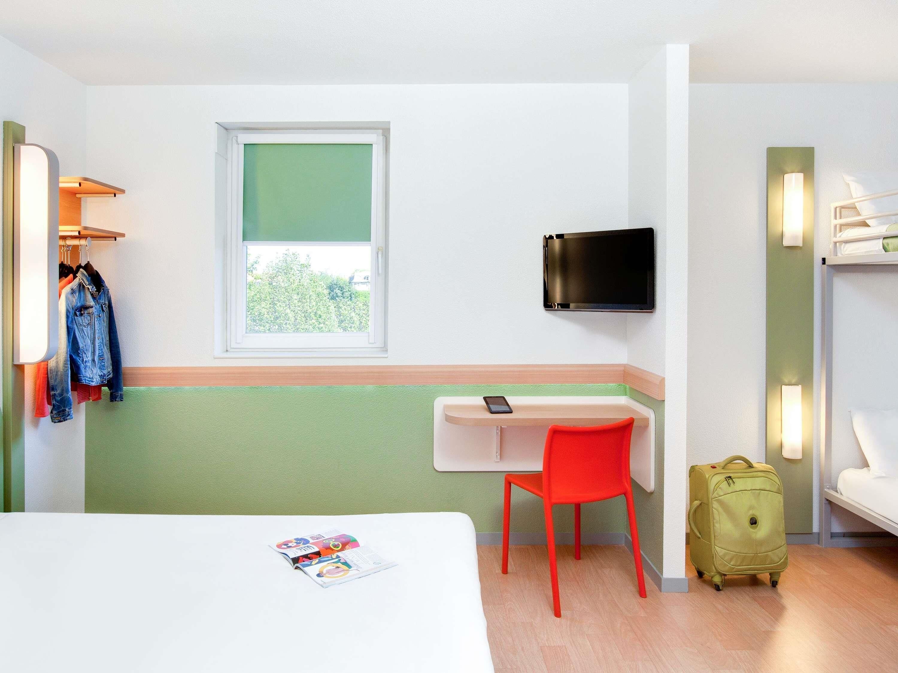 Ein Hotelzimmer mit einem Bett, einem Schreibtisch mit Stuhl, einem an der Wand montierten Fernseher und einem Koffer auf dem Boden.