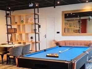 Une table de billard recouverte d’un drap bleu, un coin salon avec des sièges confortables et une étagère contenant divers objets.