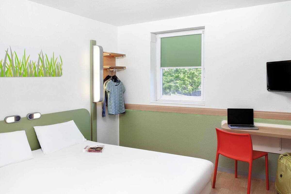 Habitación de hotel con cama, escritorio con computadora portátil, silla roja, maleta verde y ventana con vista a árboles.