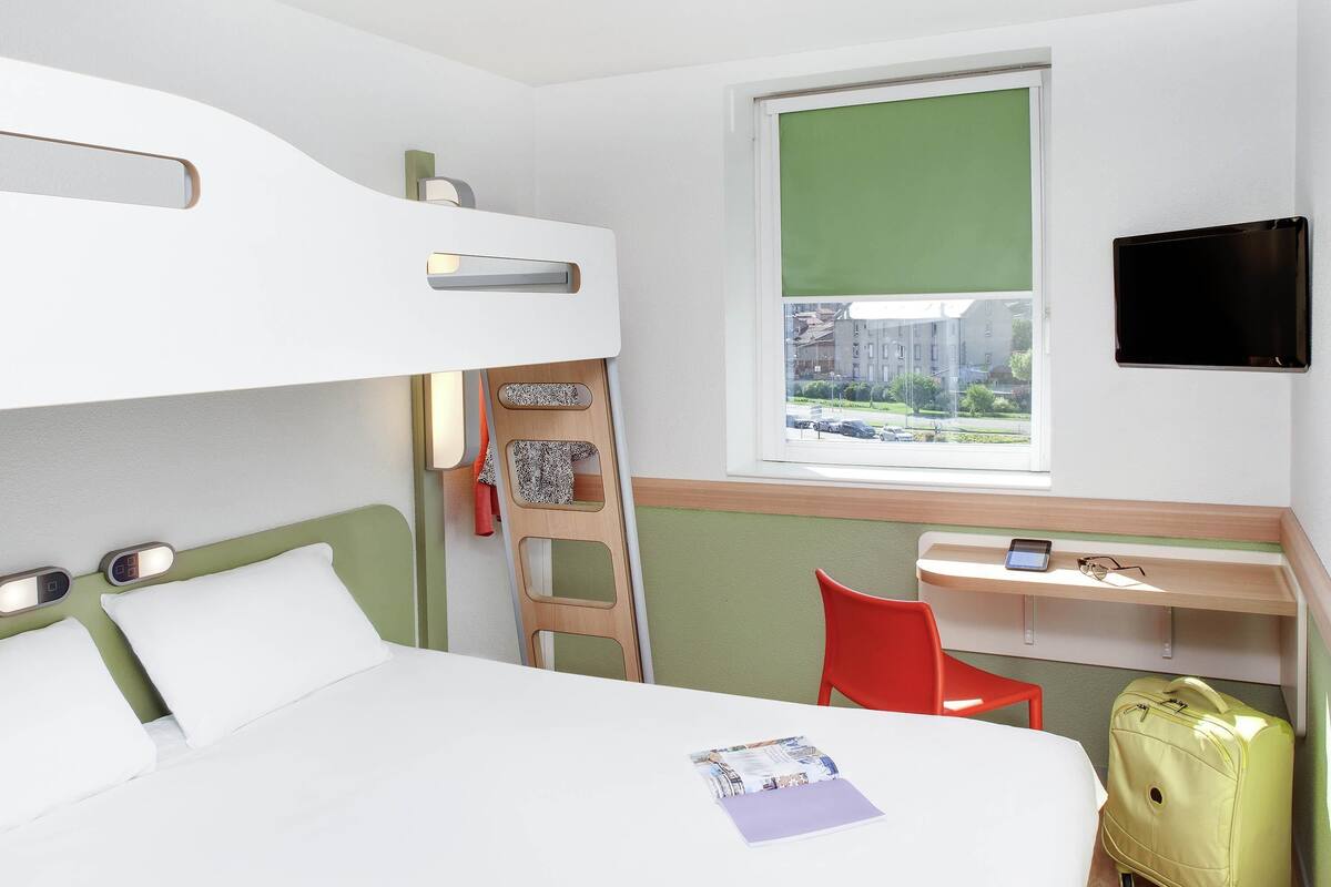 Une chambre d’hôtel avec un lit superposé, un bureau avec une chaise, une valise et un téléviseur fixé au mur.