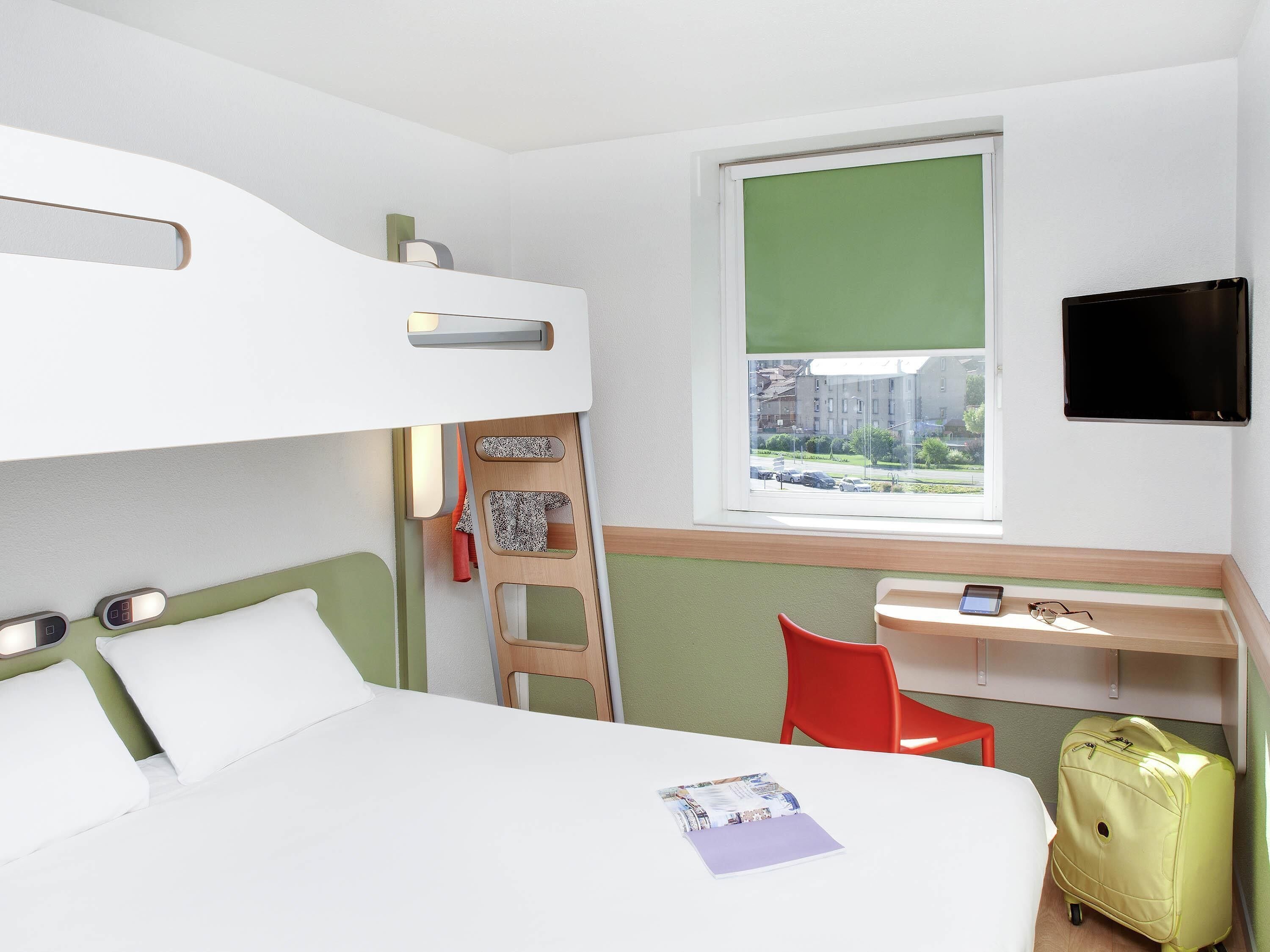 A hotel room with a bunk bed, a desk with a chair, a suitcase, and a TV on the wall.