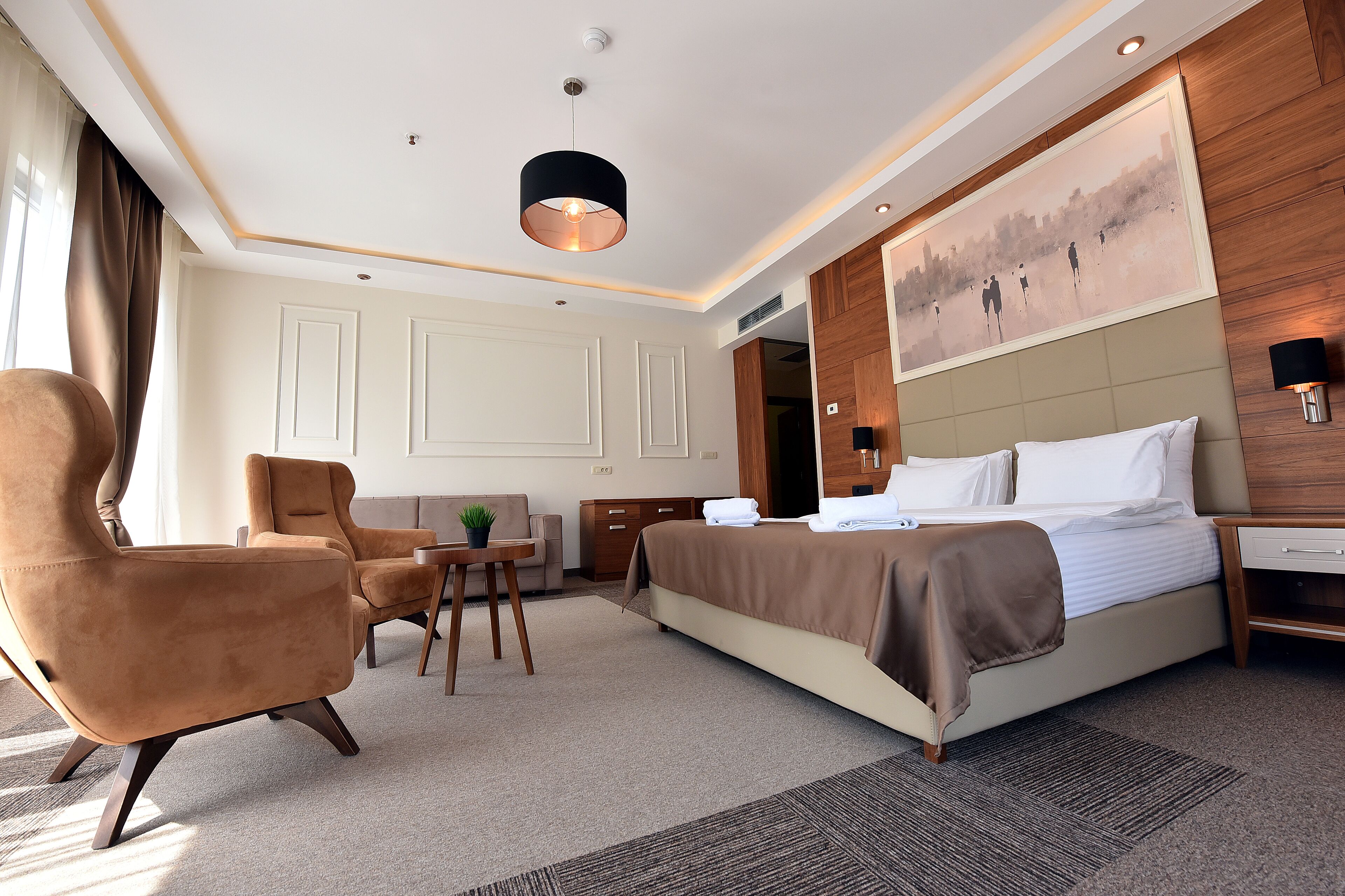 A modern hotel room with a large bed, a seating area with two chairs, a small table, and a painting on the wall.