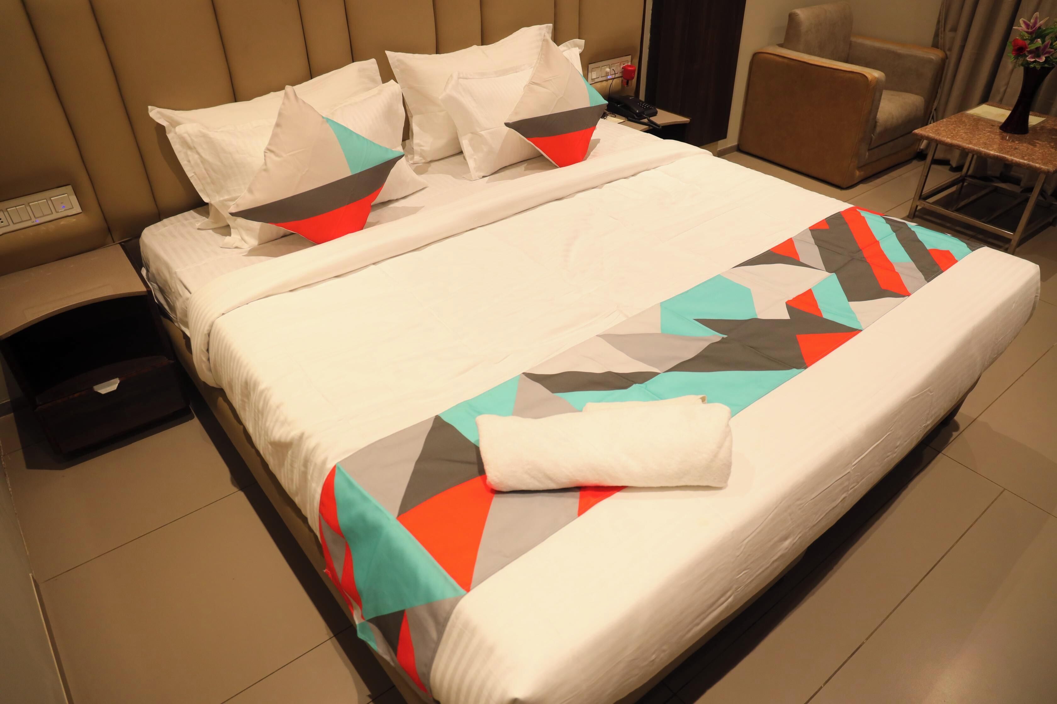 Una cama bien hecha con colcha de estampado geométrico, una mesita de noche con un florero y una silla en una habitación de hotel.