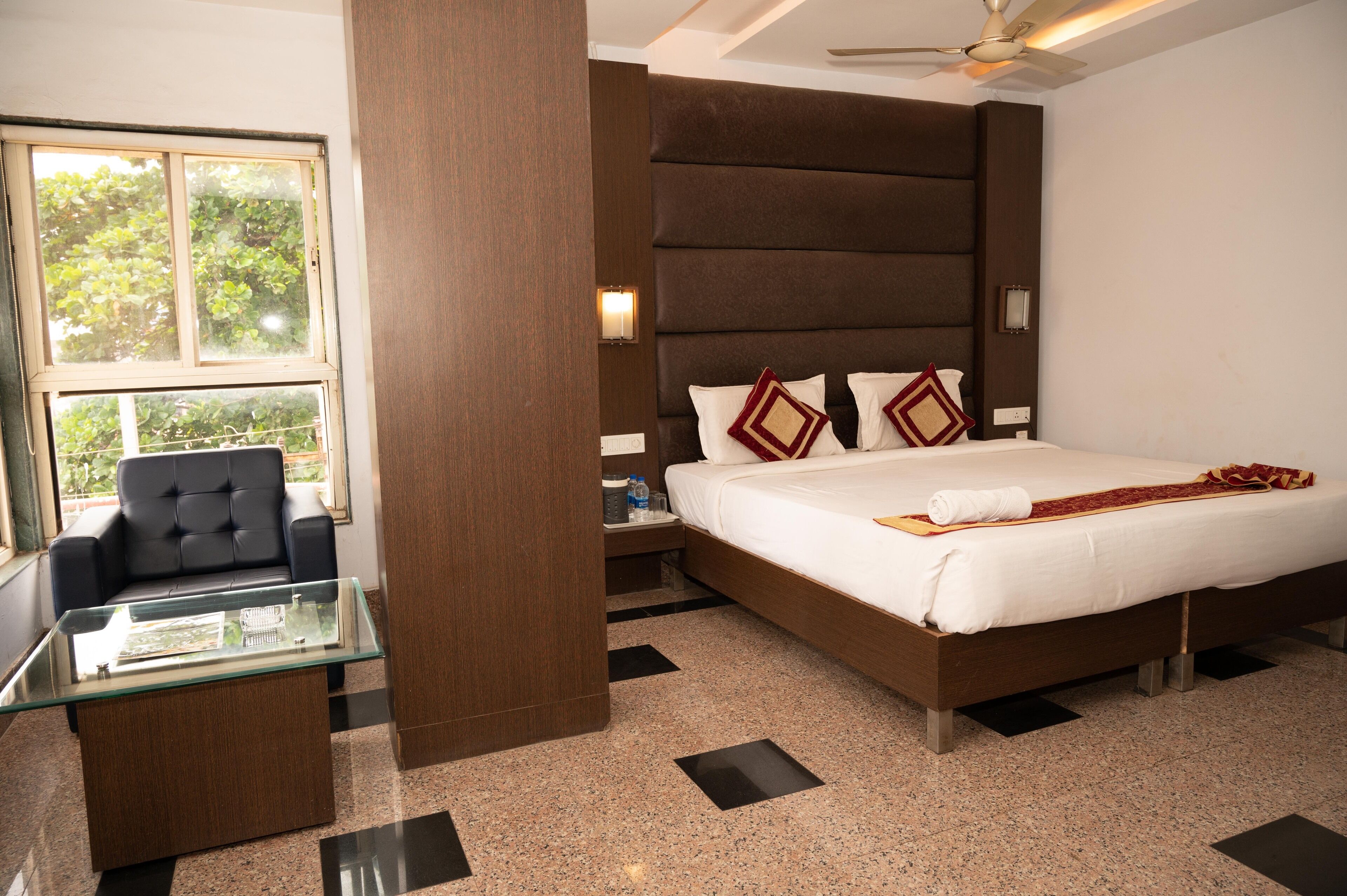 Habitación de hotel con una cama, una mesa de centro de vidrio, un sillón de cuero negro y vistas a árboles por la ventana.
